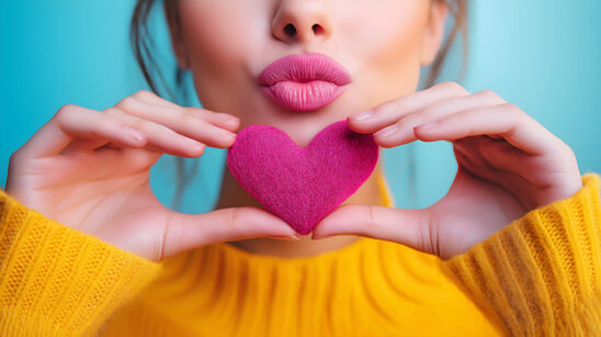 Close-up of a beautiful woman face with heart shaped hands in front of her mouth which forms a kiss. Love Sign.